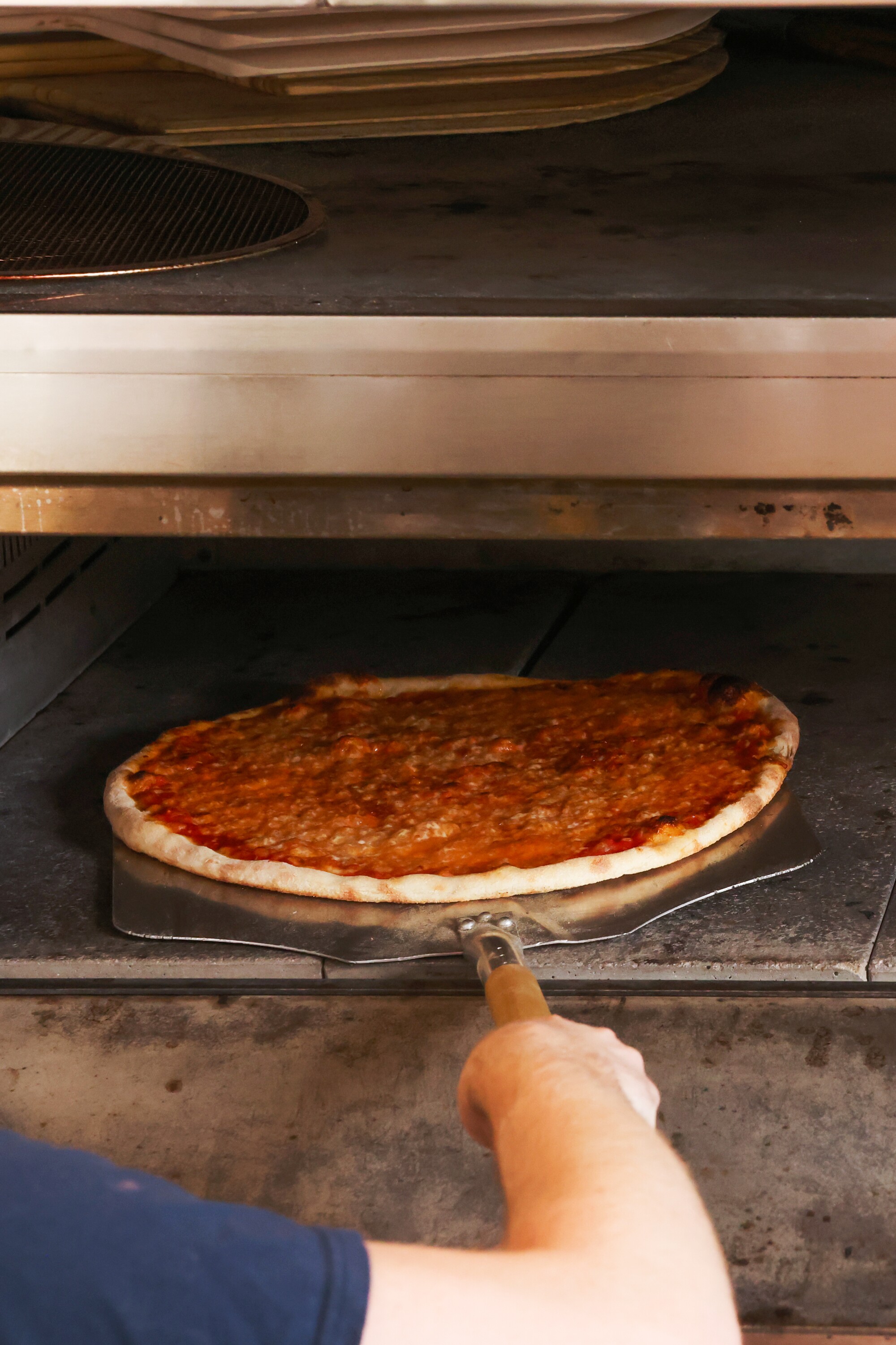 A hand uses a pizza peel to retrieve a cheese pizza from a pizza oven.