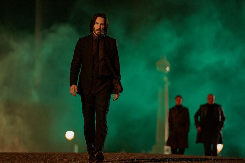 Keanu Reeves walking toward the camera in a black suit amid a haze of green fog