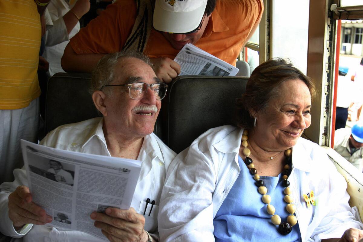 Gabriel Garcia Marquez sits in a train carriage alongside his wife as she smiles and looks out the window. 
