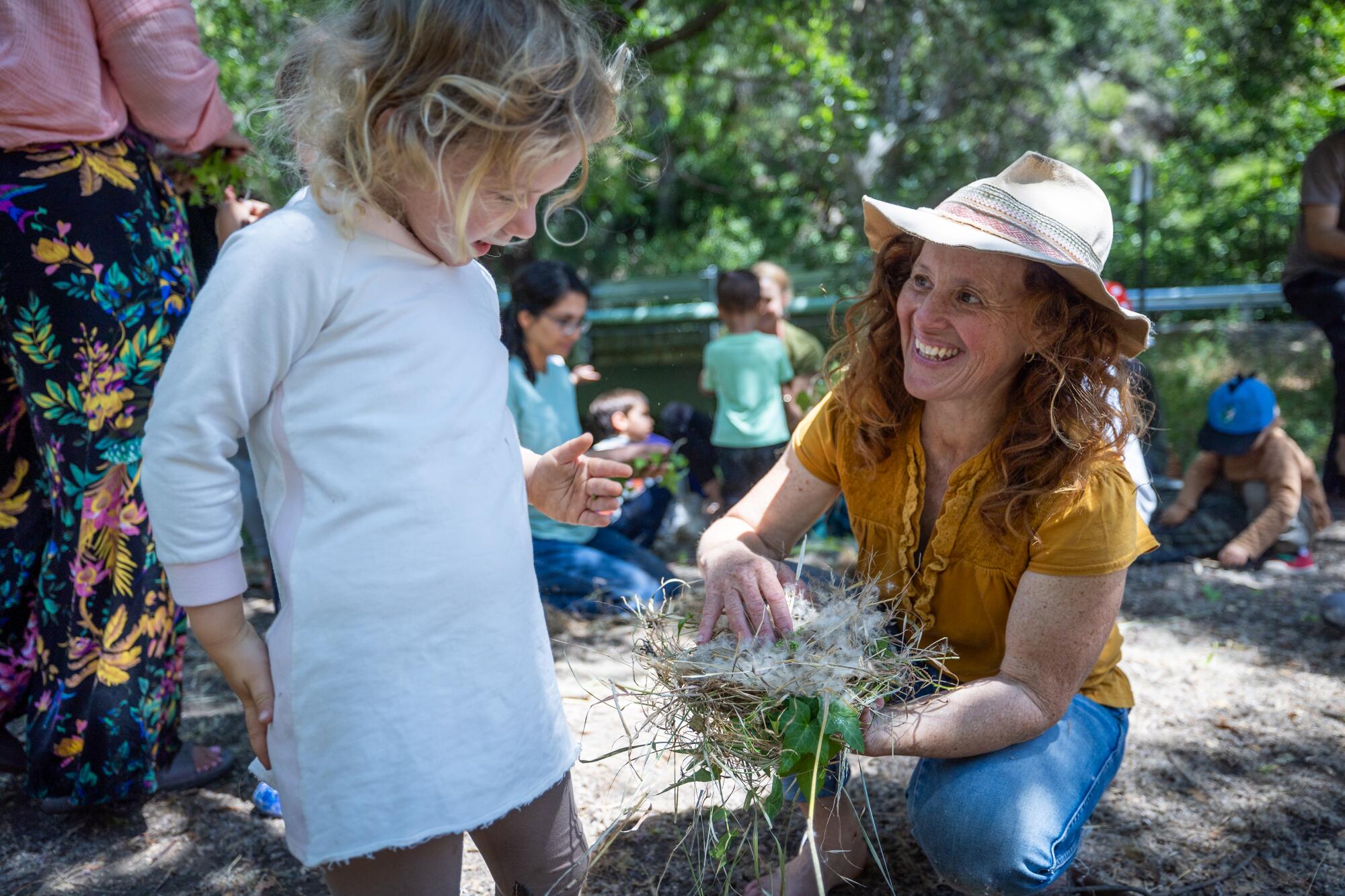 A woman shows a child a handful of twigs and grass.