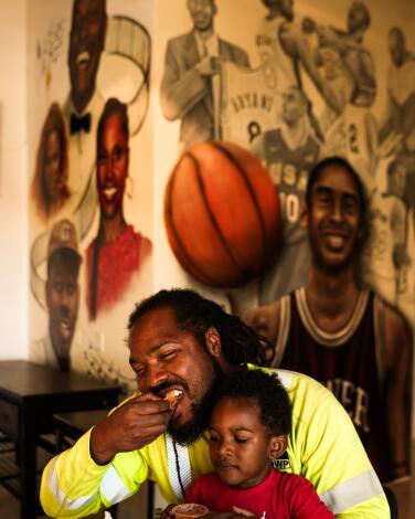 A man eats tacos and holds a young boy in front of a mural featuring basketball players.