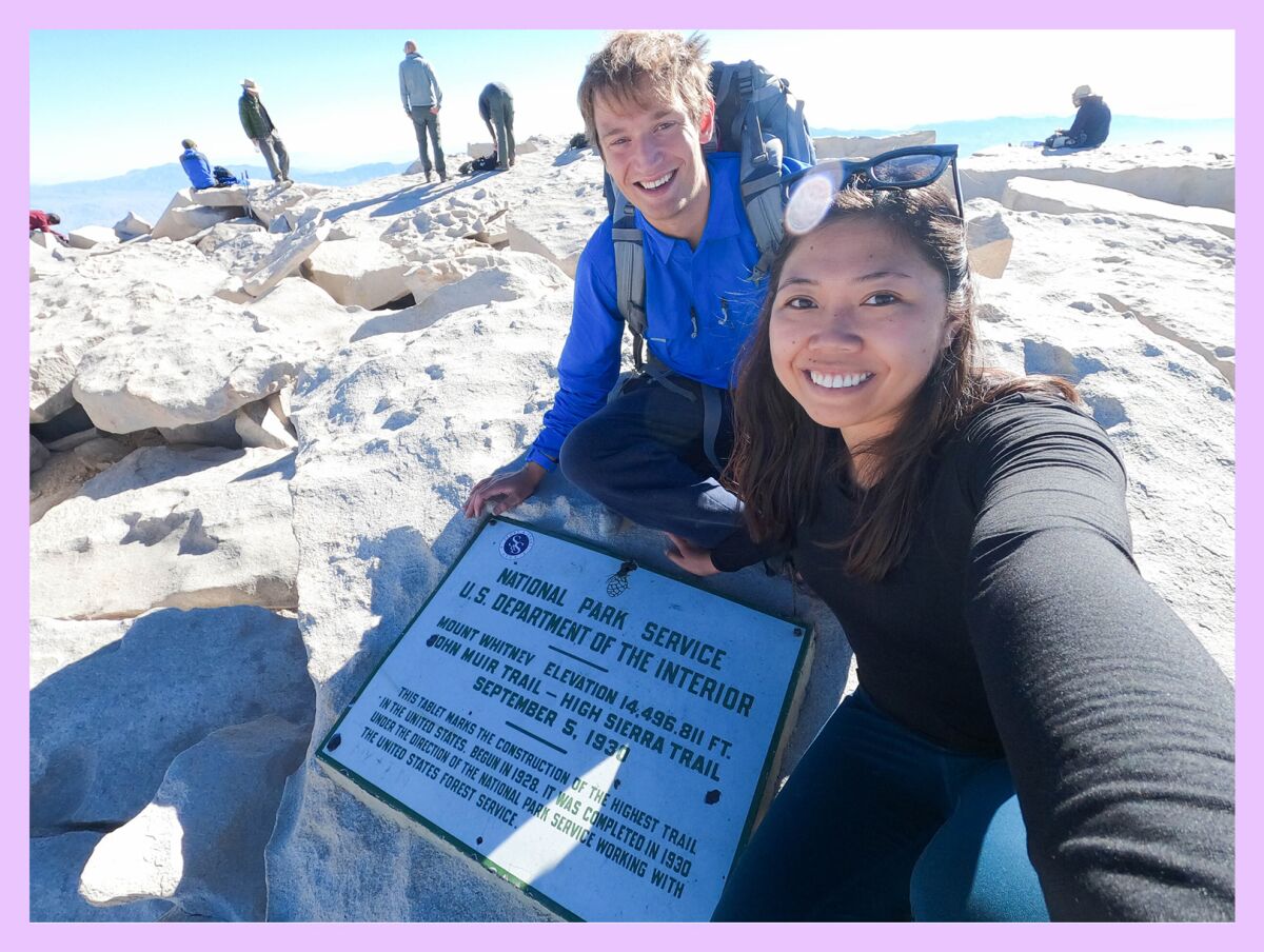 man and a woman smile at a plaque at the top of a mountain