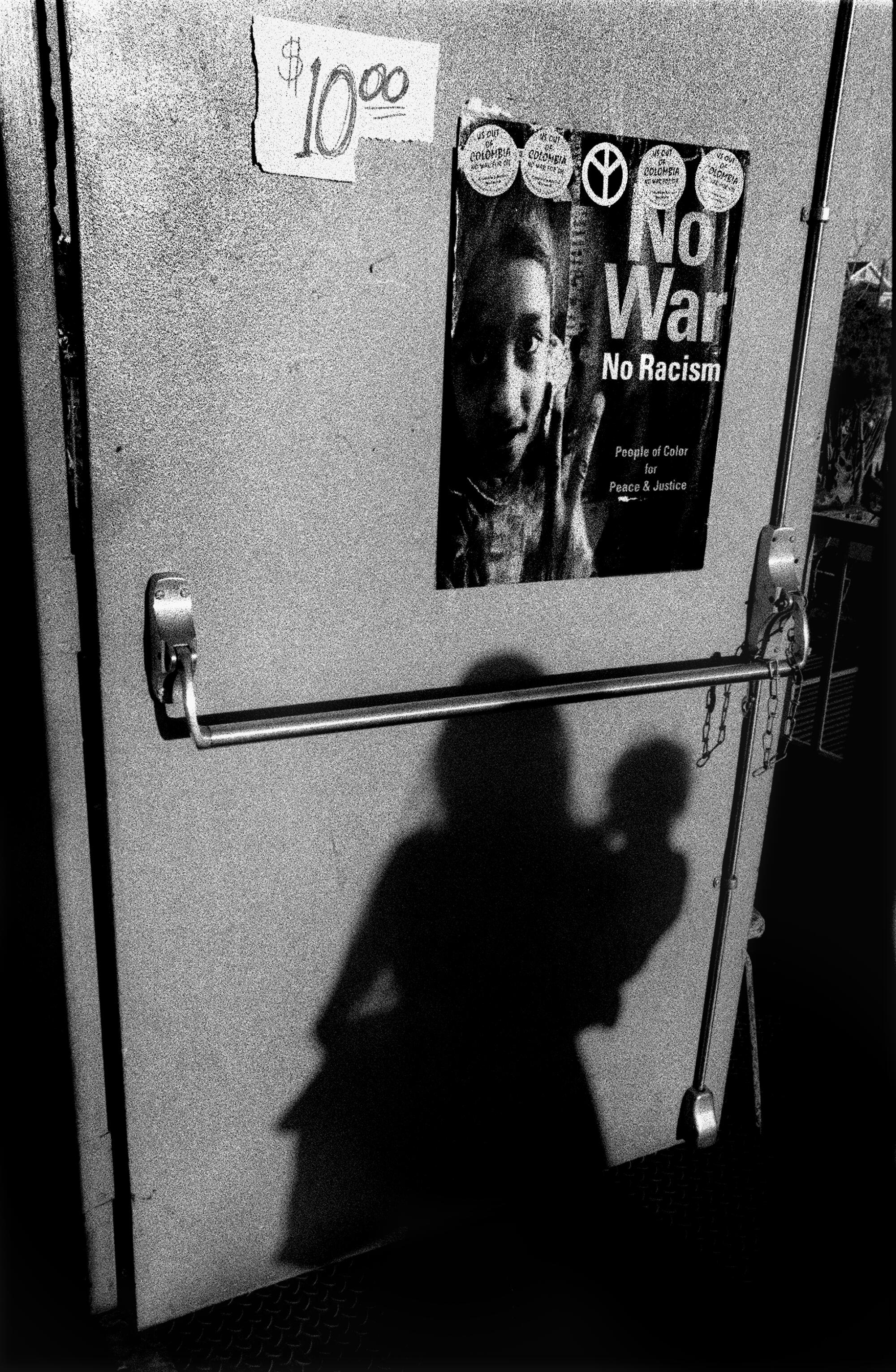 The shadow of a woman carrying a child is seen against a door with a poster that reads "No War, No Racism."