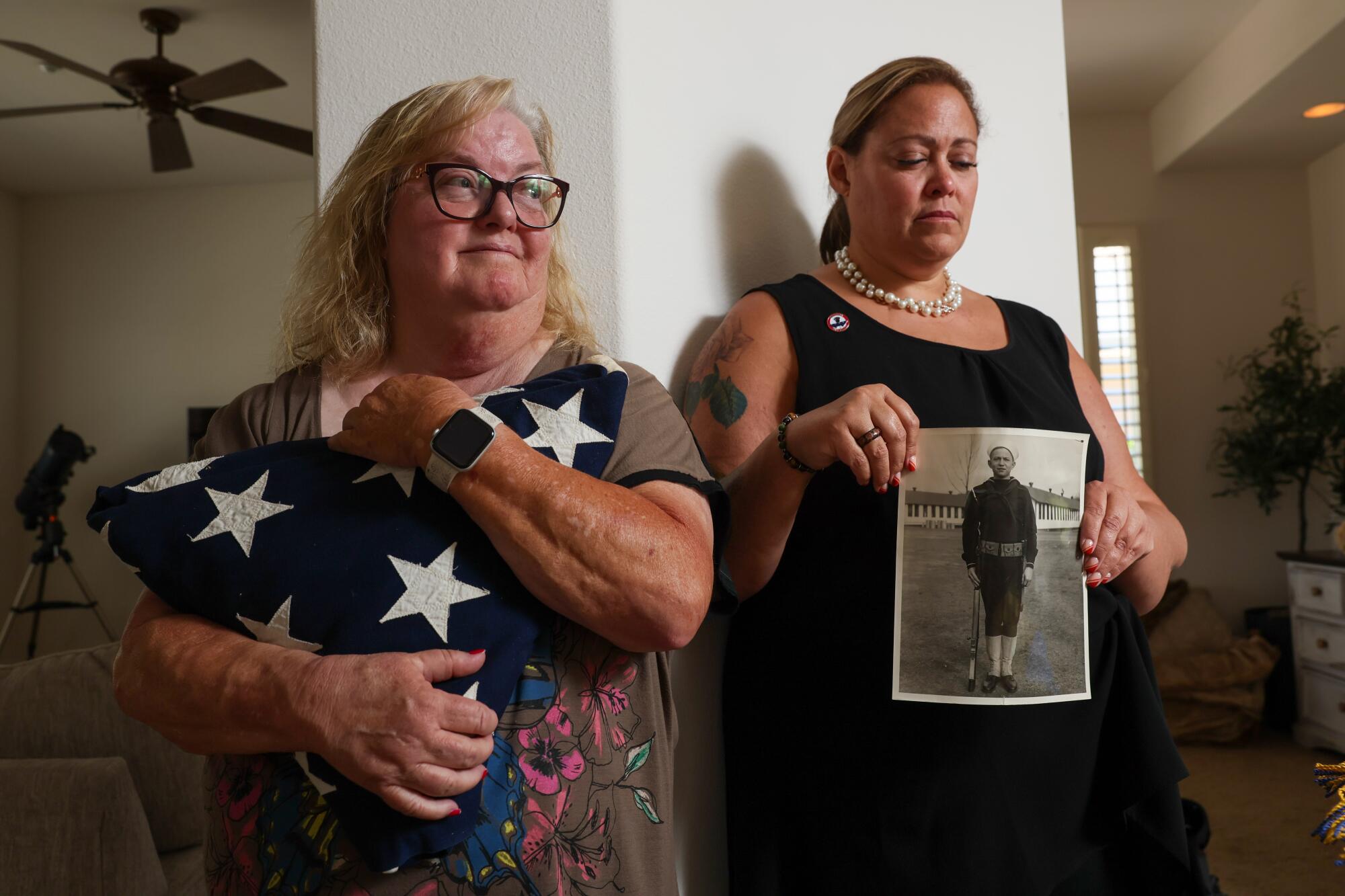 A woman with blond hair and glasses, holding a folded flag, left, stands next to a woman holding a black-and-white photograph