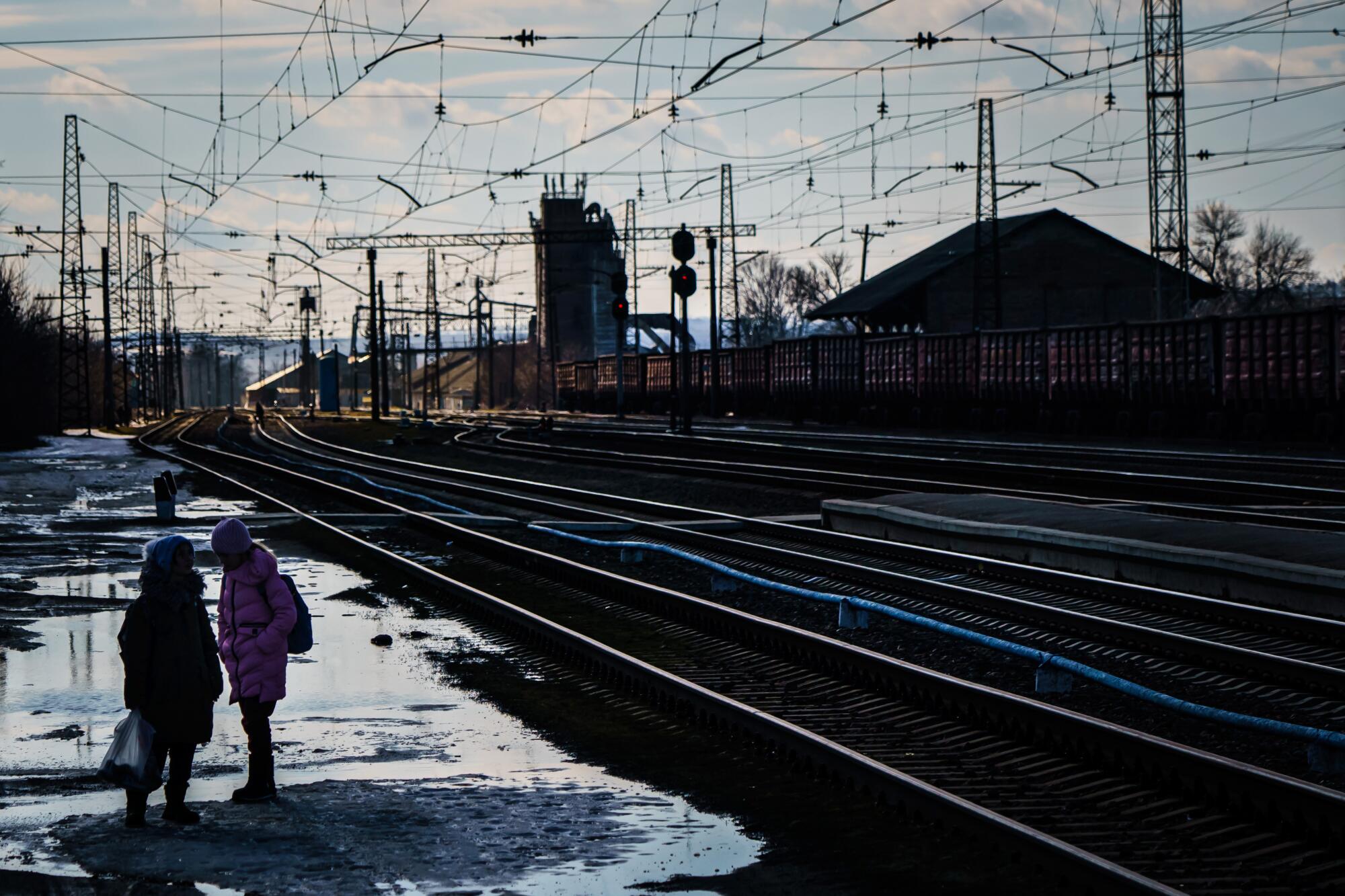 Two people stand near rail tracks amid rain puddles outside at a train station.