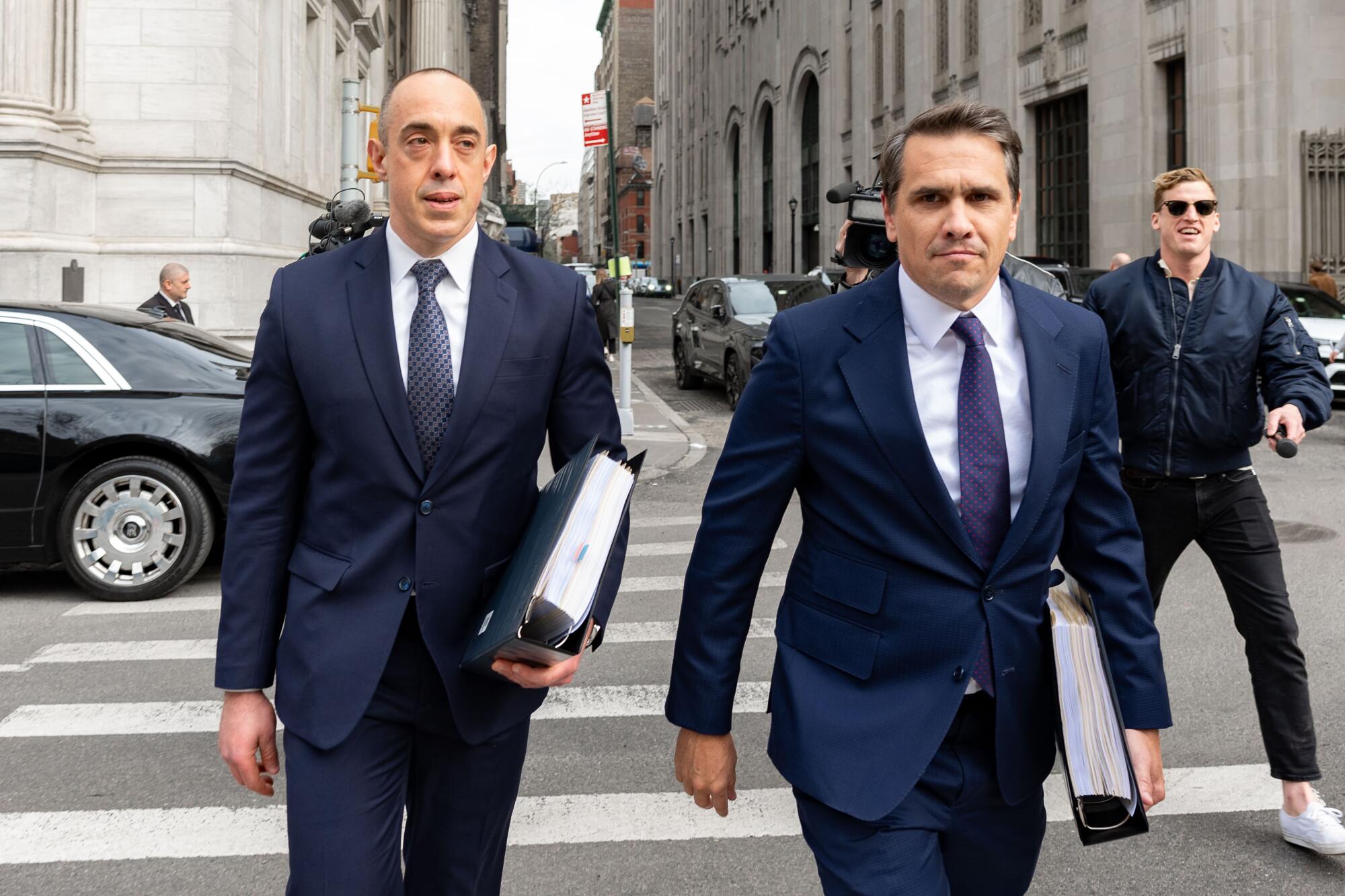 Dois homens em ternos azuis com pastas grossas atravessam uma rua da cidade, enquanto um homem segurando um microfone tenta alcançá-los.