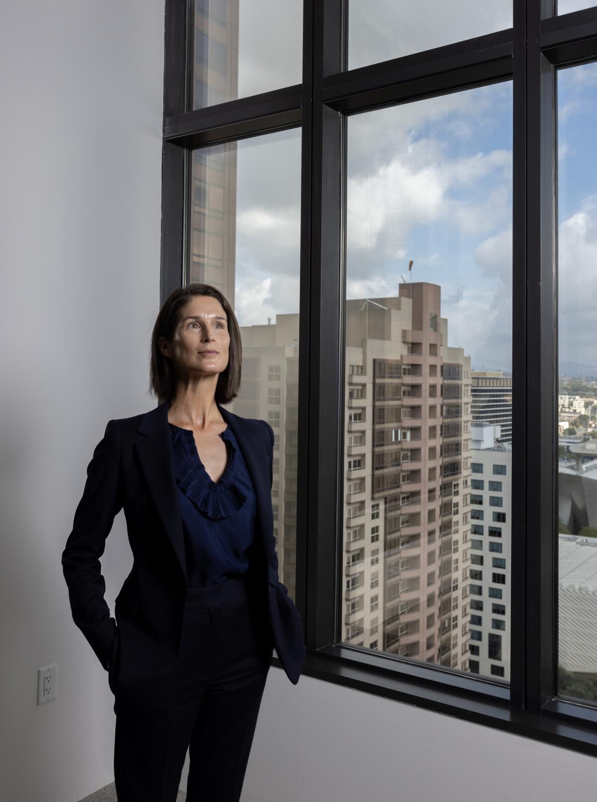 A woman standing in a white room, next to a large window with a city view of tall buildings below