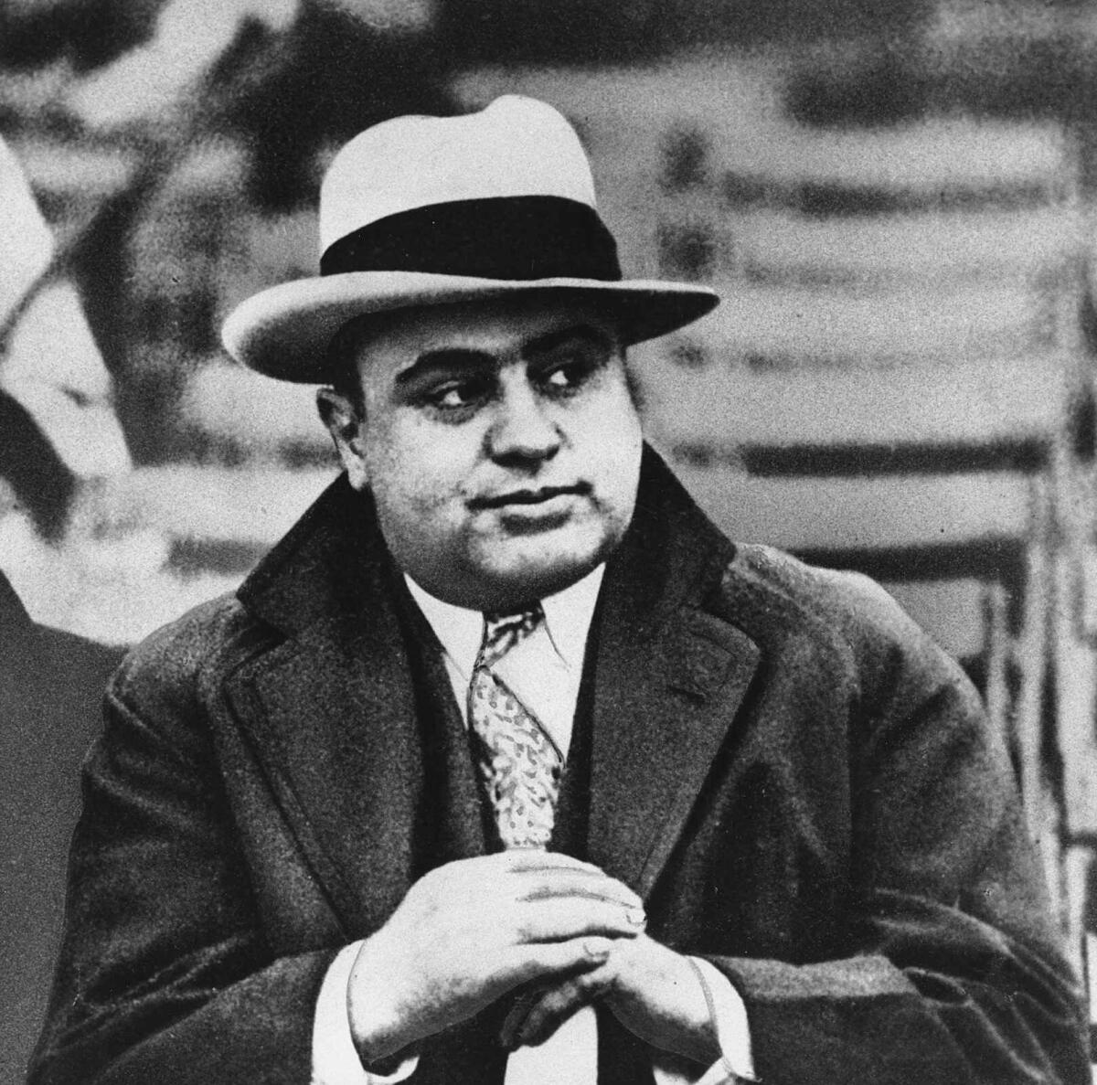 A man in tie and winter coat and fedora sits in bleachers.