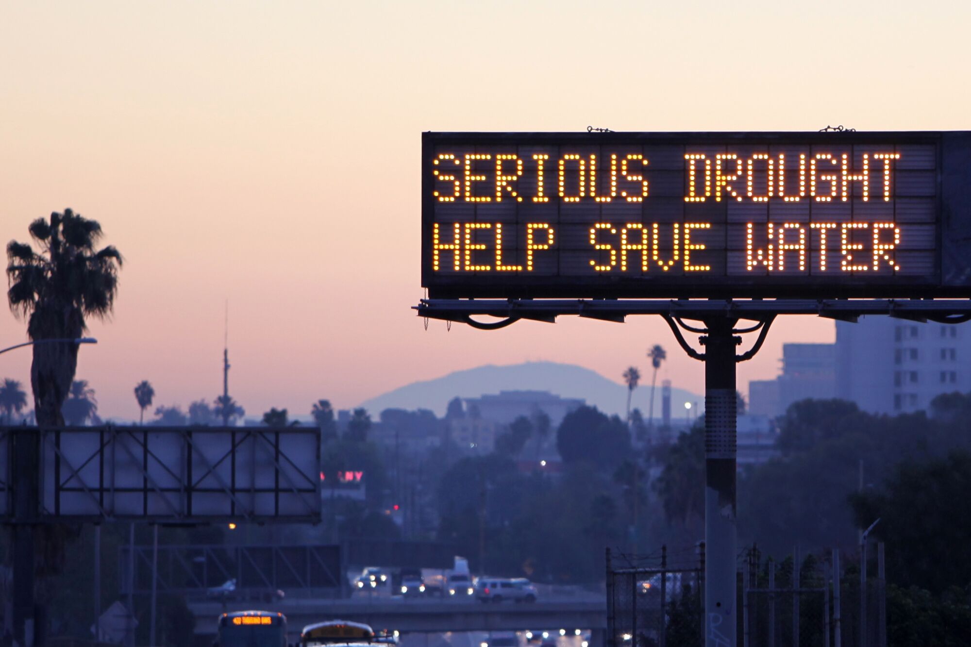 A Los Angeles freeway at dusk is capped by an illuminated sign that reads: "Serious Drought, Help Save Water"