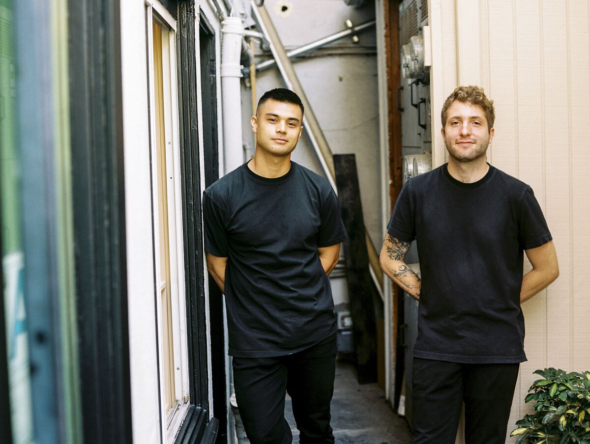 Two young men dressed all in black stand between buildings. 