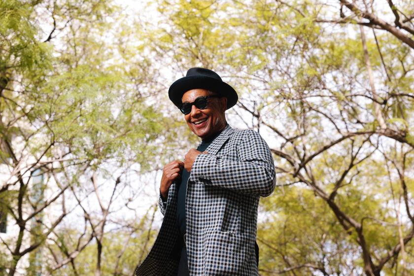 Giancarlo Esposito tugging at the lapels of a black-and-white blazer, wearing a black hat and dark glasses in front of trees