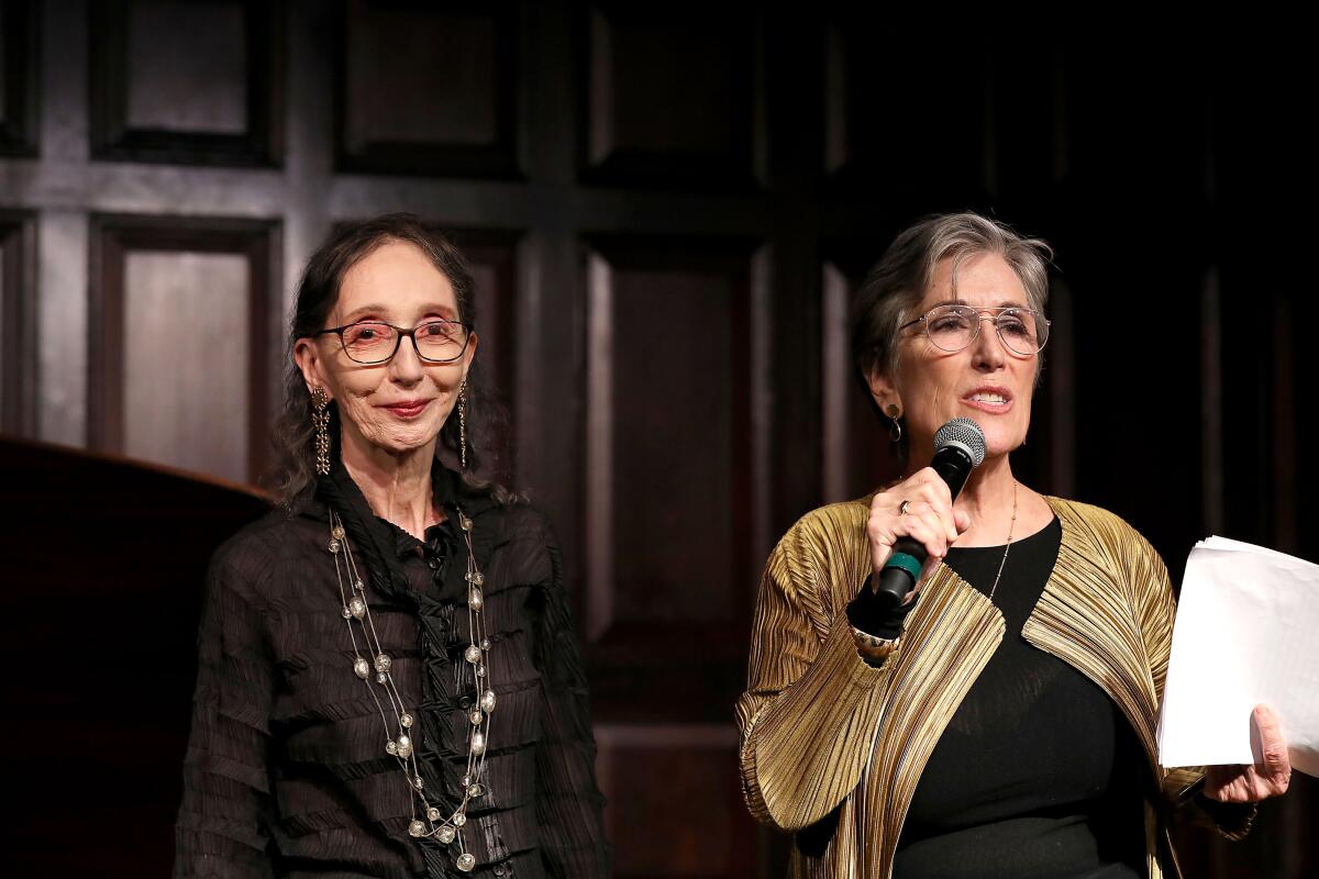 Joyce Carol Oates, in a dark shirt, stands on stage next to Cedering Fox, in a gold jacket, at a literary event
