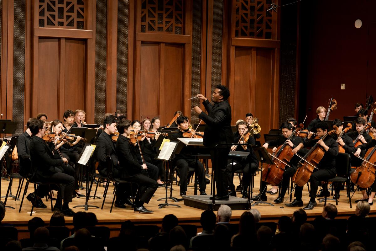 A wood-paneled stage is filled with musicians conducted by Anthony Parnther.