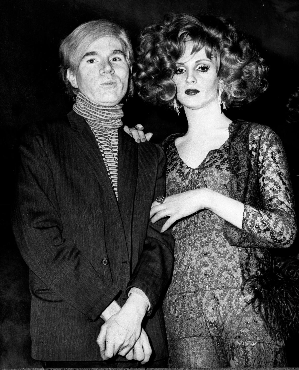 Andy Warhol, in a turtleneck and suit, stands next to Candy Darling, with short curly hair and a lace dress.