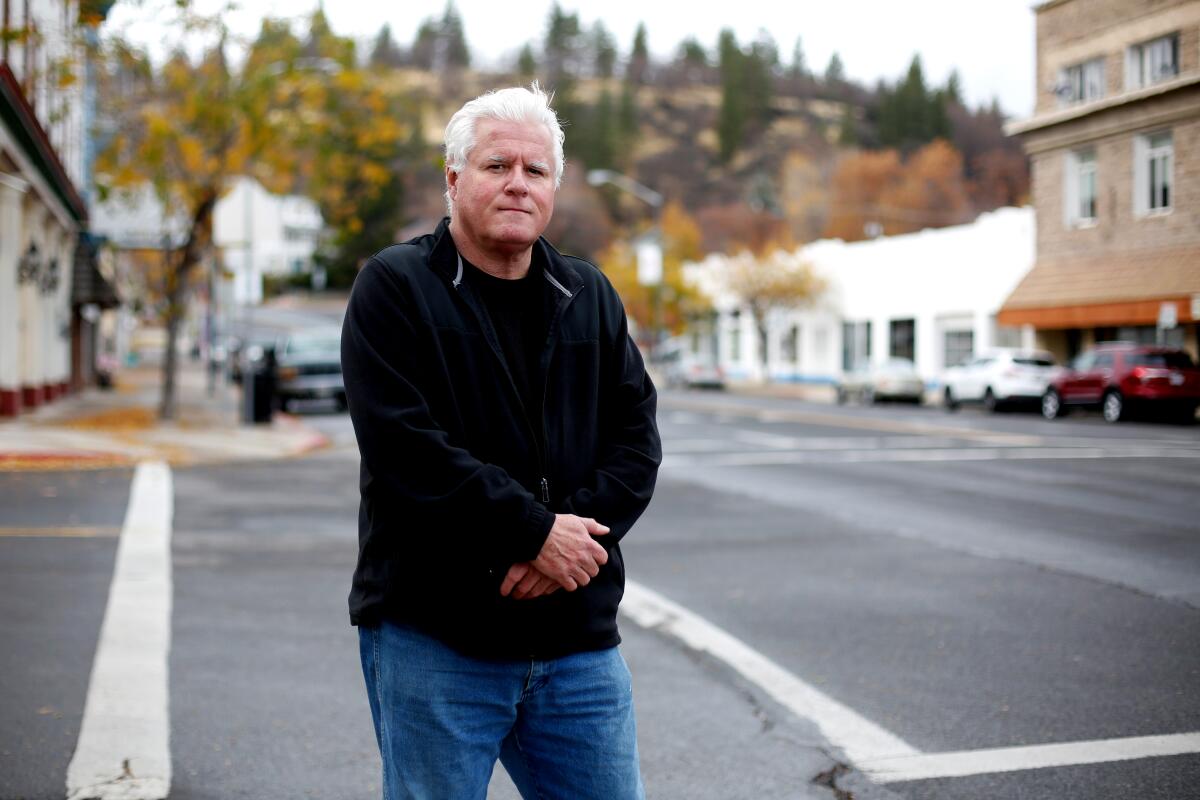 A man stands on a downtown street with a hill full of trees in the distance.