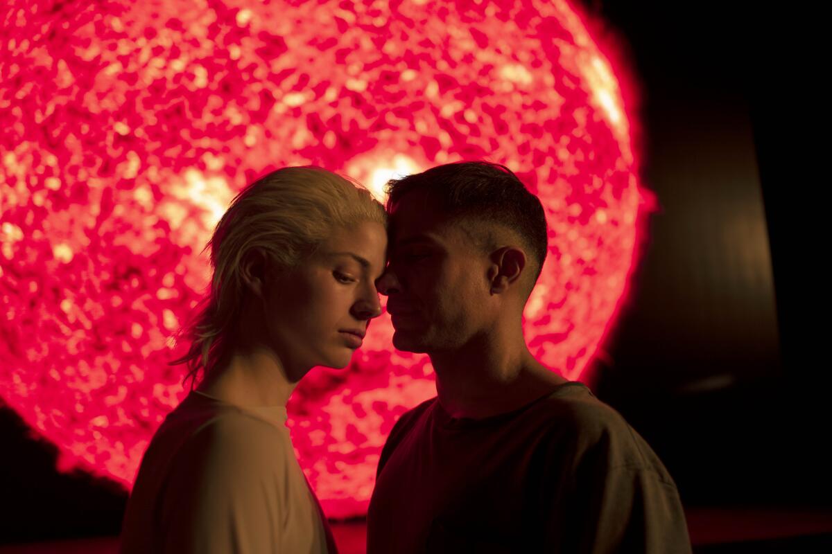 A woman and a man lean their heads together as they stand before a glowing red ball.