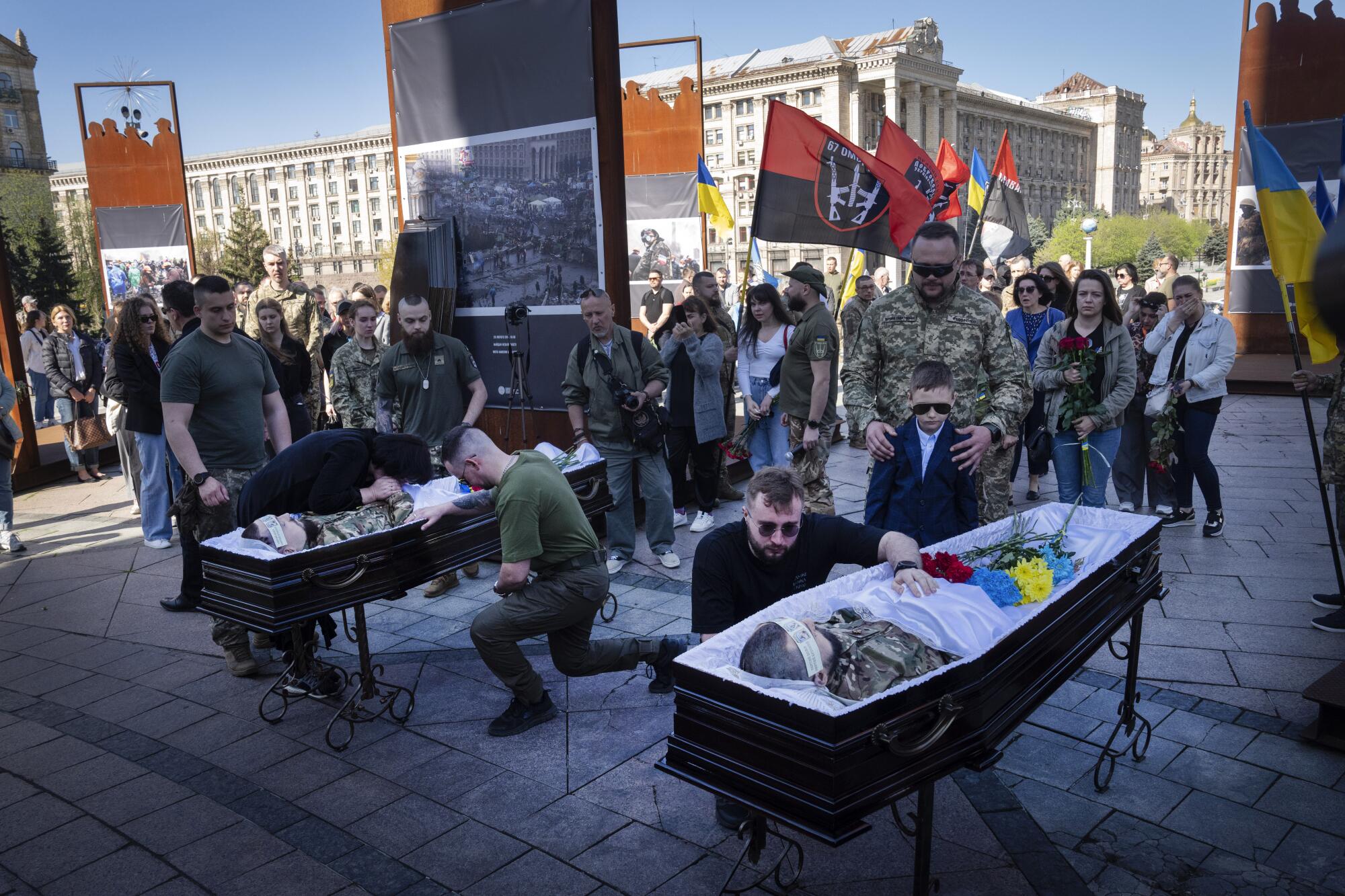 People stand and kneel by the bodies of two men in their coffins, with red-and-black flags nearby