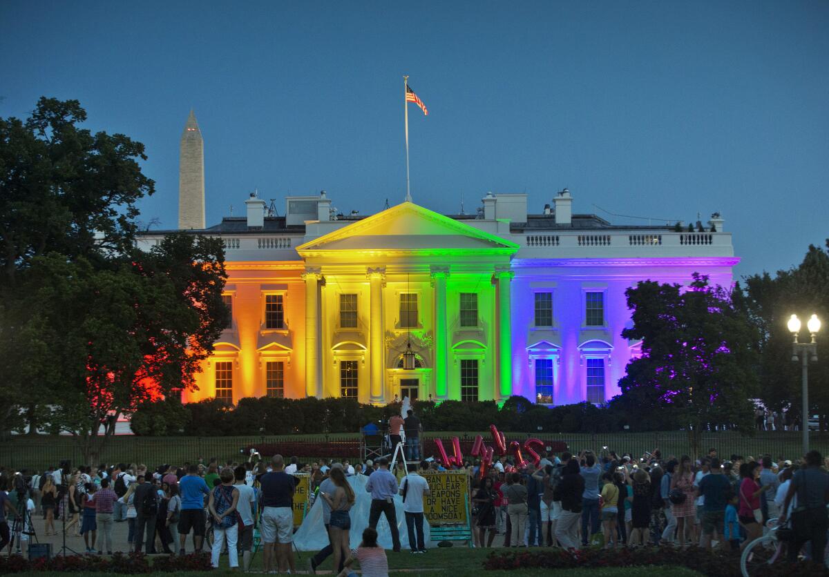 The White House is lit up in rainbow colors of orange, yellow, green, blue and purple. A crowd is gathered In the foreground.