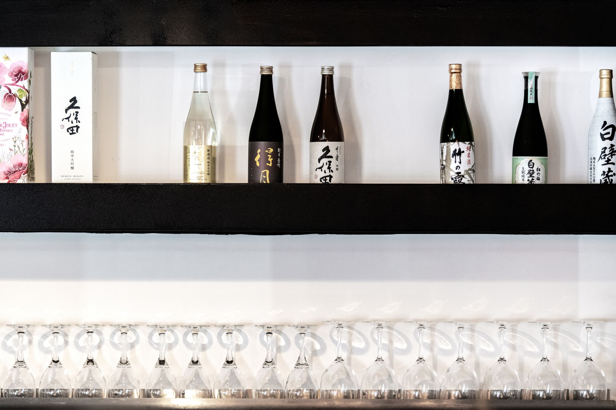 A straight-on view of black shelves on a white wall, lined with drinking glasses and bottles of sake.