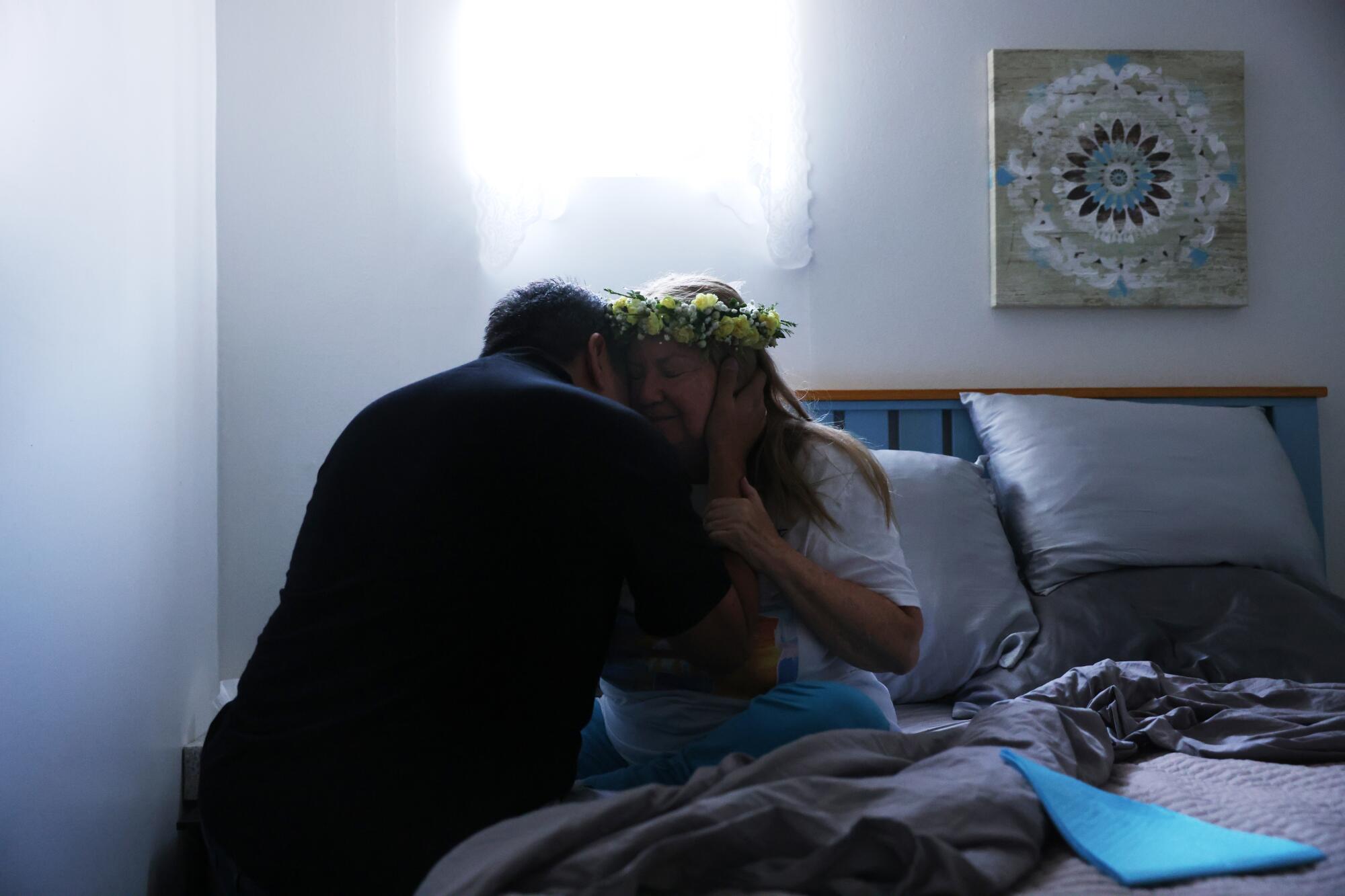 A person hugs a woman in a flower crown sitting in bed