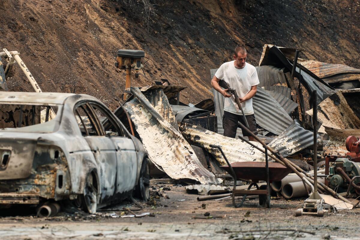 A man holds a rifle while standing among burned out structures and a burned out car..