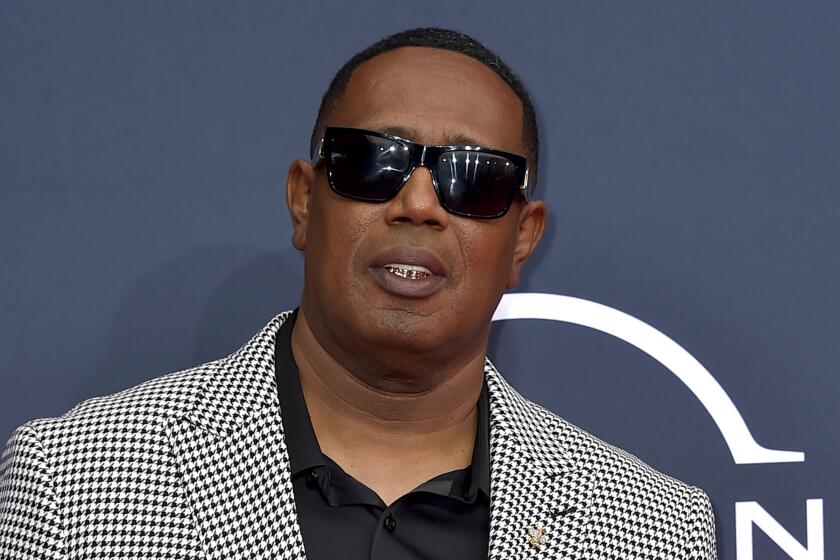 Master P poses in sunglasses and a checkered suit against a gray background.