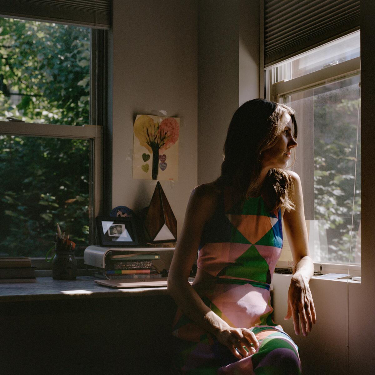 Leslie Jamison, in a colorful geometric-print dress, sits and looks out a window.