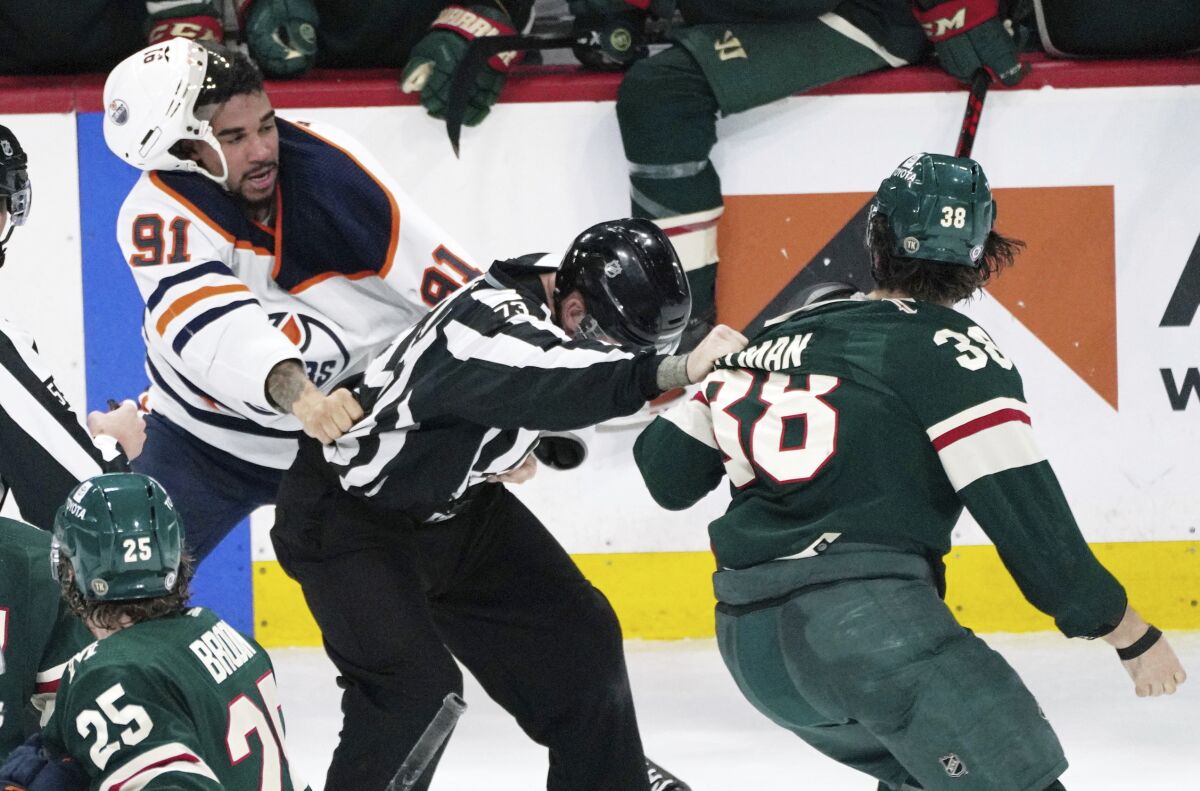 An official, center, gets caught between Edmonton Oilers' Evander Kane (91) and Minnesota Wild's Ryan Hartman (38) during a skirmish in the third period of an NHL hockey game Tuesday, April 12, 2022, in St. Paul, Minn. Both players received game misconduct penalties. The Wild won 5-1, with Hartman scoring two of the goals. (AP Photo/Jim Mone)