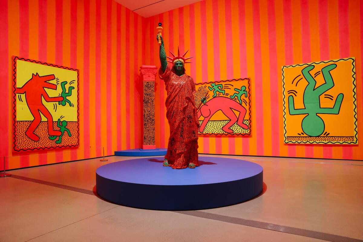A gallery covered in striped wallpaper bears colorful paintings by Keith Haring, as well as a Statue of Liberty statue