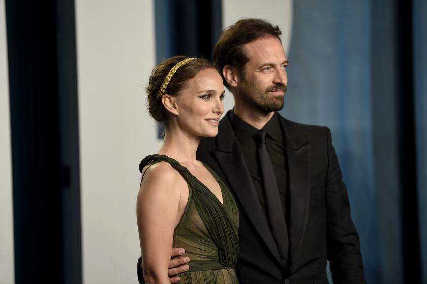 Natalie Portman with a braided updo and golden headband in a sleeveless black gown next to Benjamin Millepied in a black suit