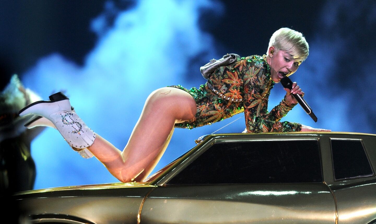 Miley Cyrus leans in atop a car, showing off her cowboy boots with a money symbol.