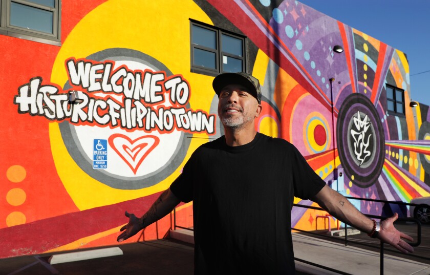A man in a black T-shirt and cap stands before a bright "Welcome to Historic Filipinotown" mural, arms spread wide.