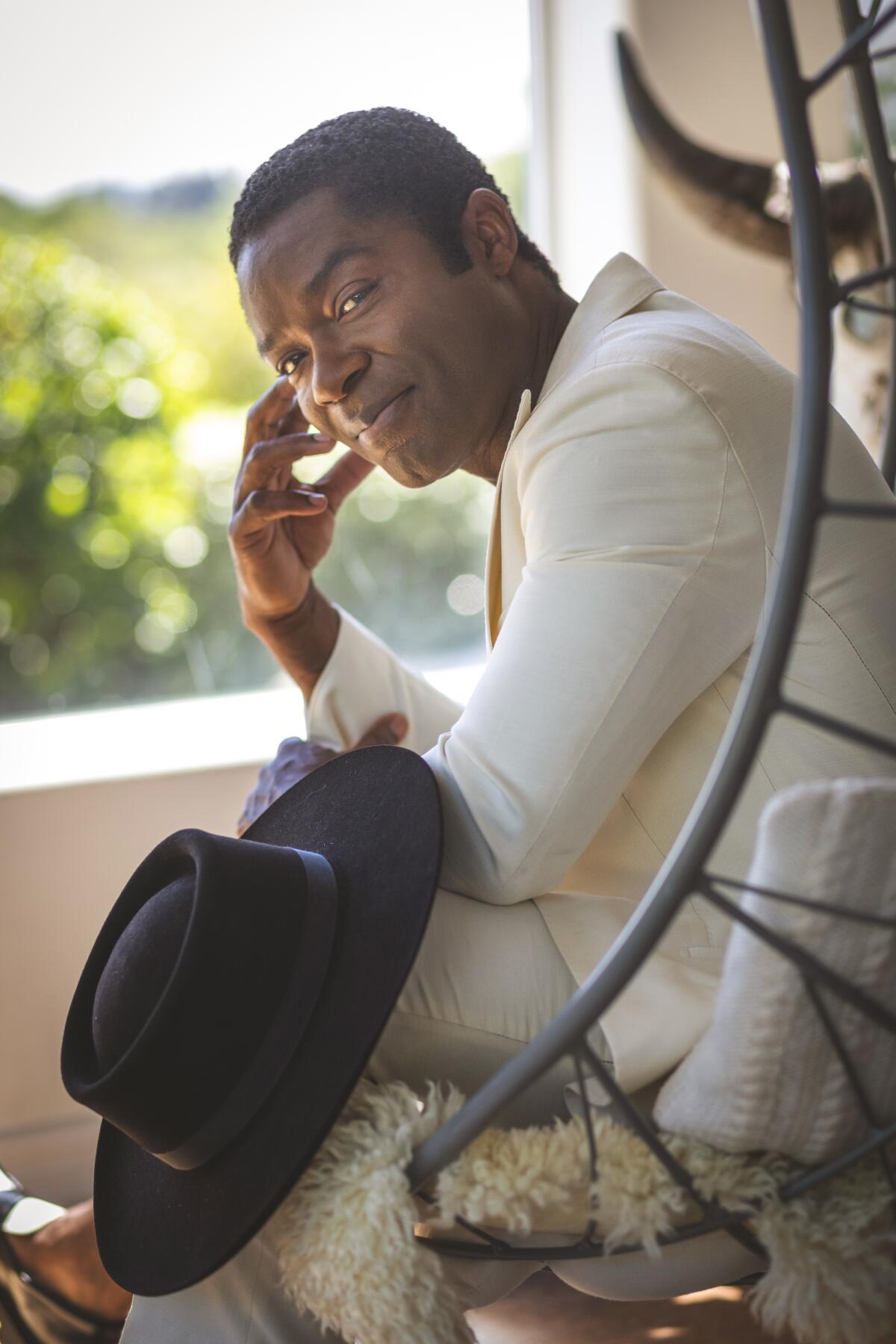 Actor David Oyelowo wears a white suit as he sits on his porch resting a black cowboy hat on his knee.