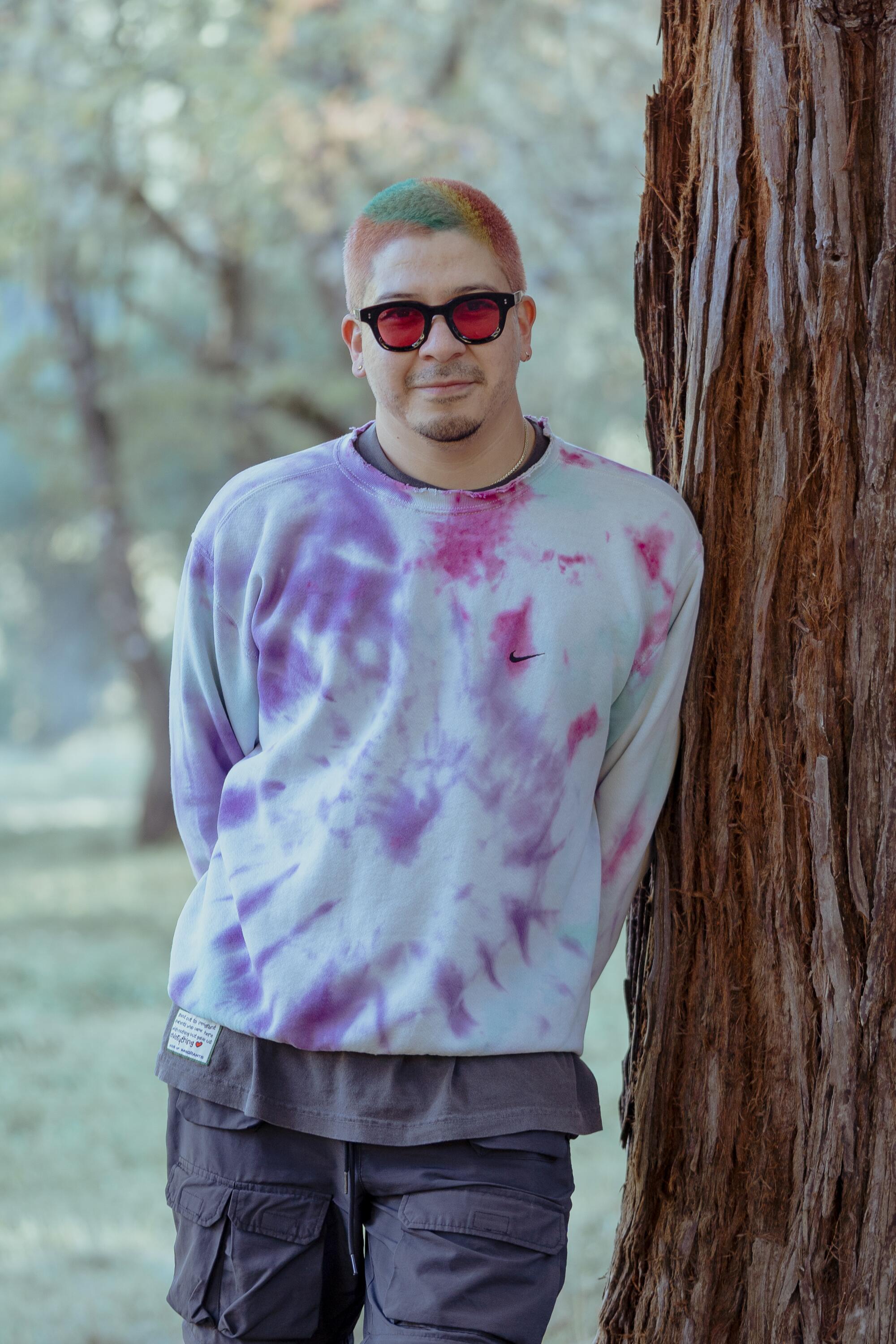 Man in tie-dye sweatshirt leans against a tree