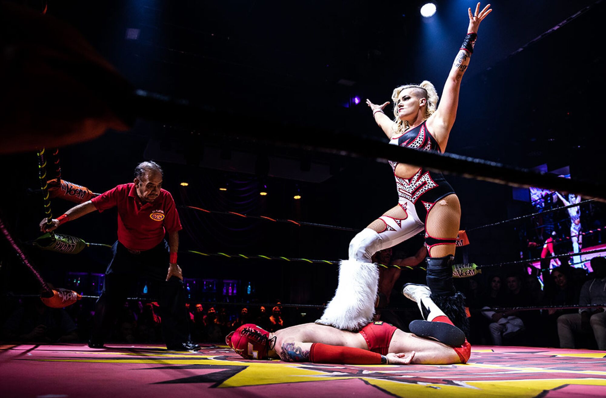 A female wrestler stands in the ring with arms raised in triumph and one foot on the back of her defeated foe