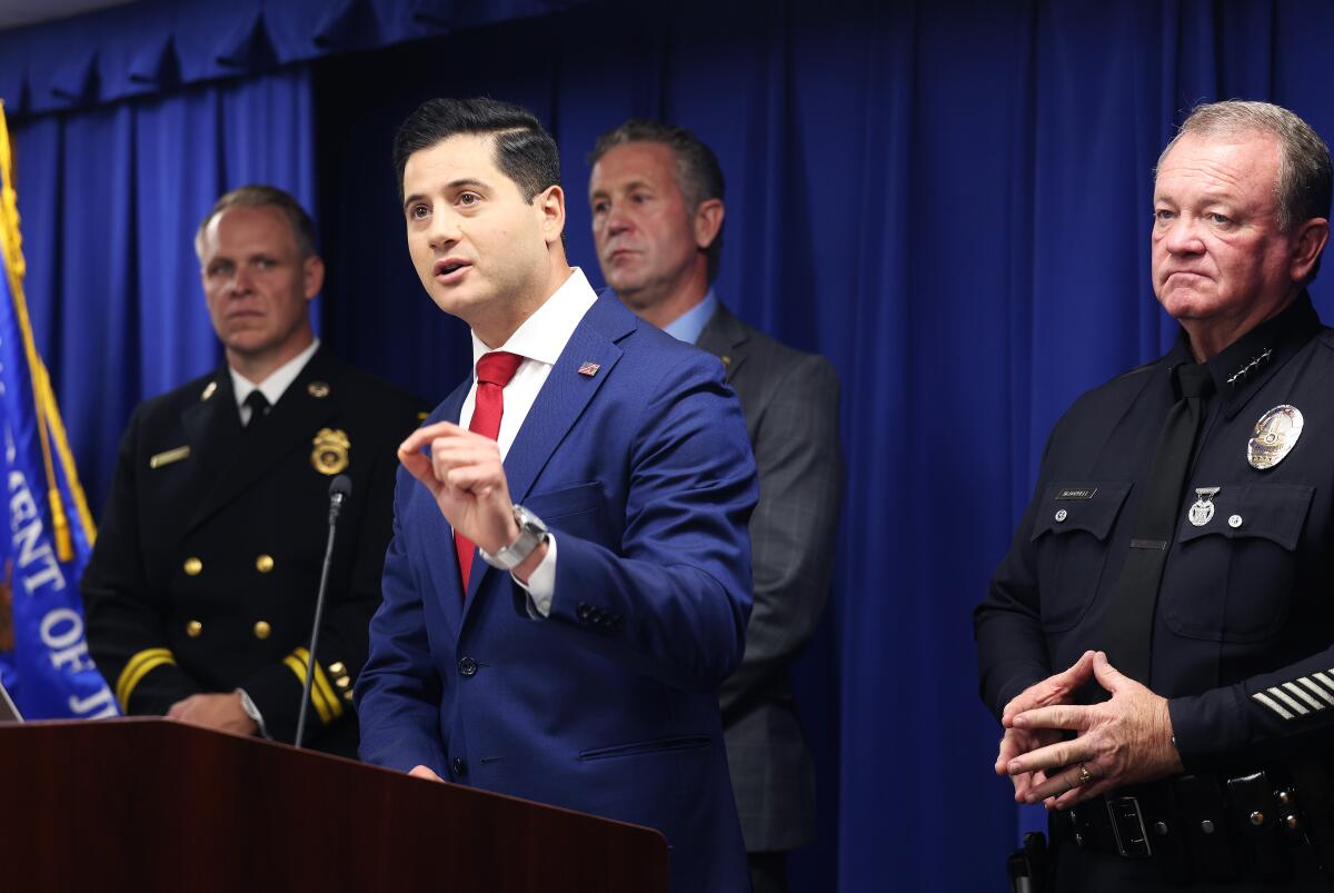 A man in a blue suit speaks into a microphone with uniformed police officers during a press conference