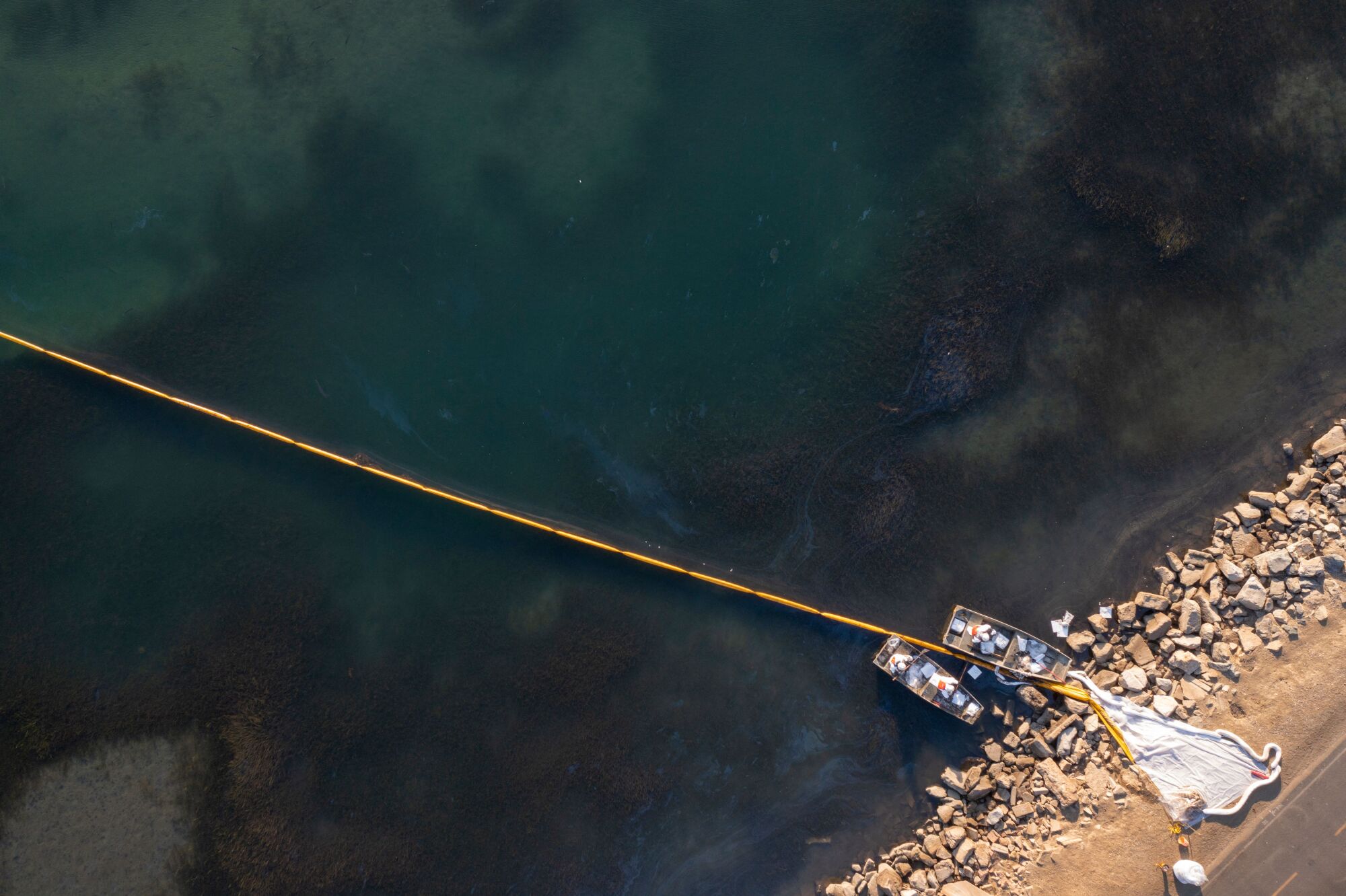 Aerial view of a line of booms stretches across water, with two boats at end, near shoreline 