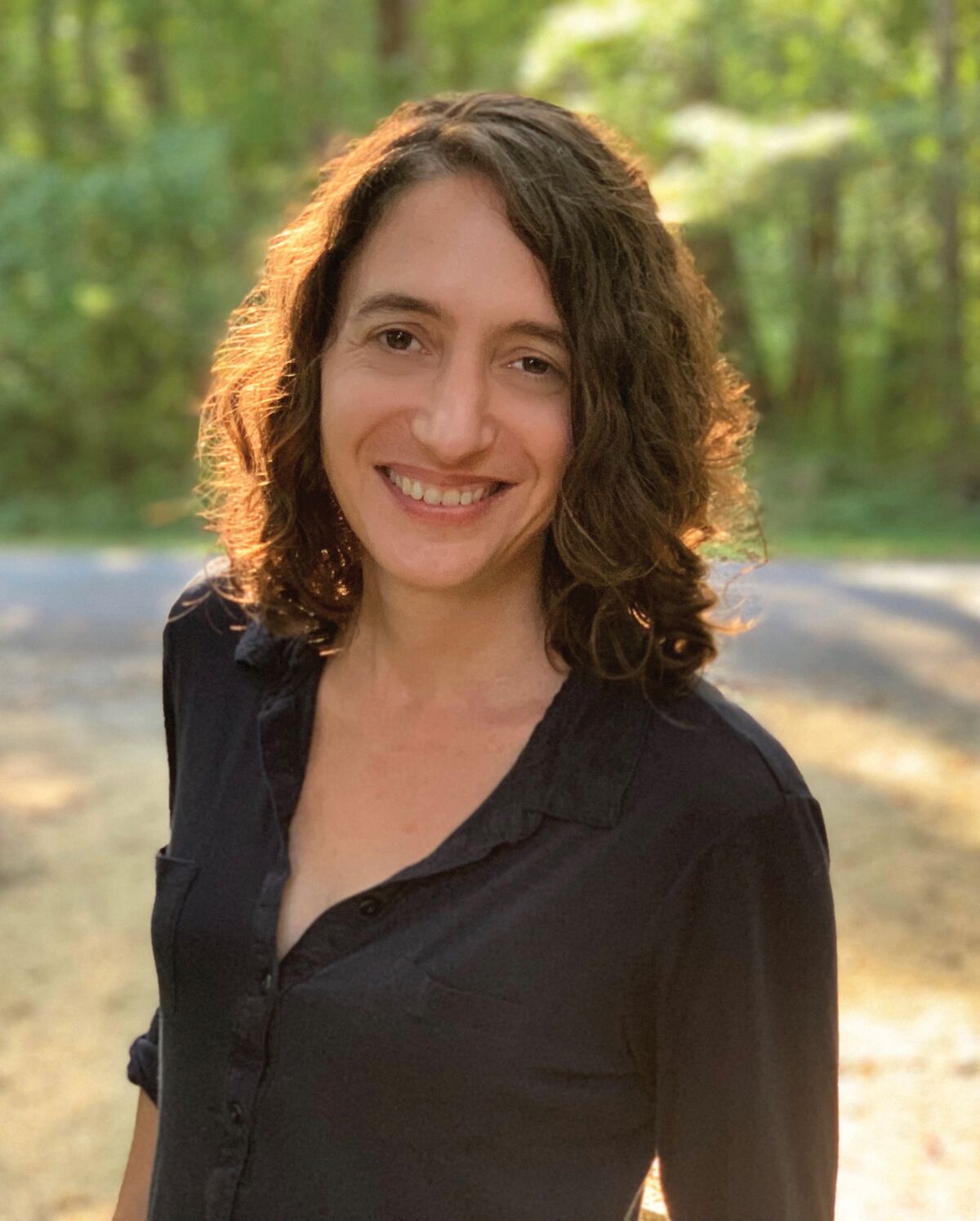 A woman with brown hair in a dark shirt stands outdoors, smiling.