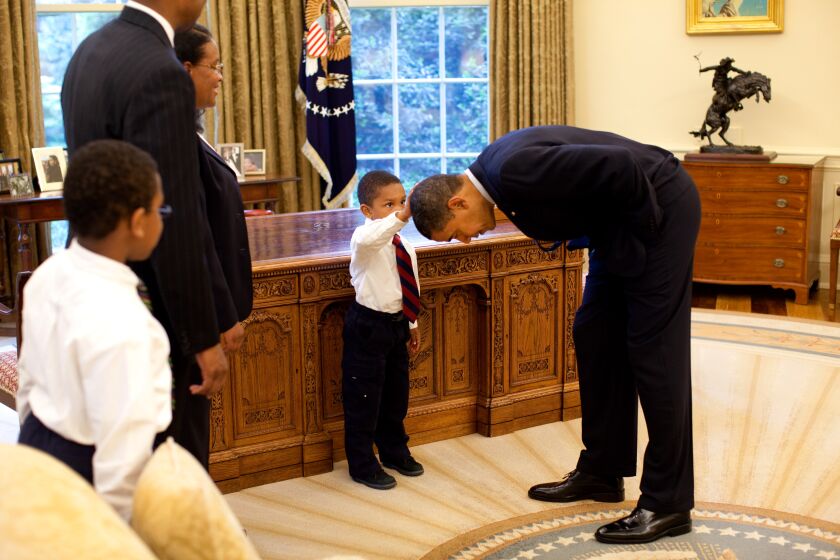 President Barack Obama bends over so the son of a White House staff member can pat his head during a visit to the Oval Office