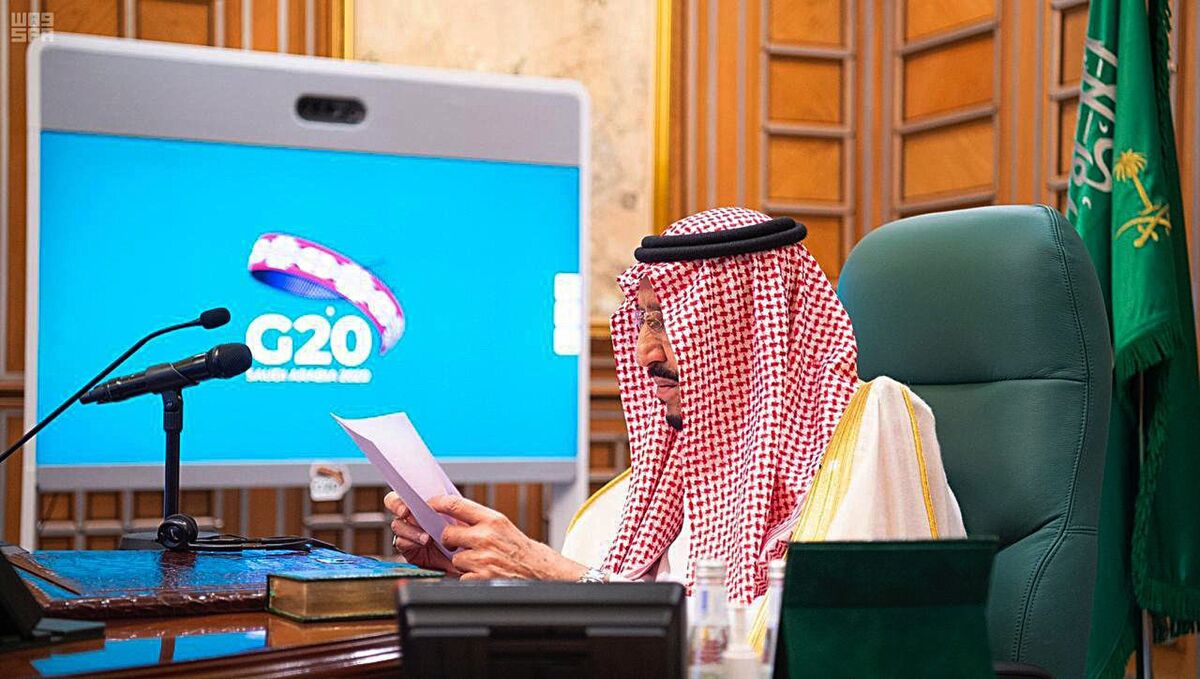 Saudi King Salman sits at a desk holding papers while having a video call with the Group of 20 logo behind him.