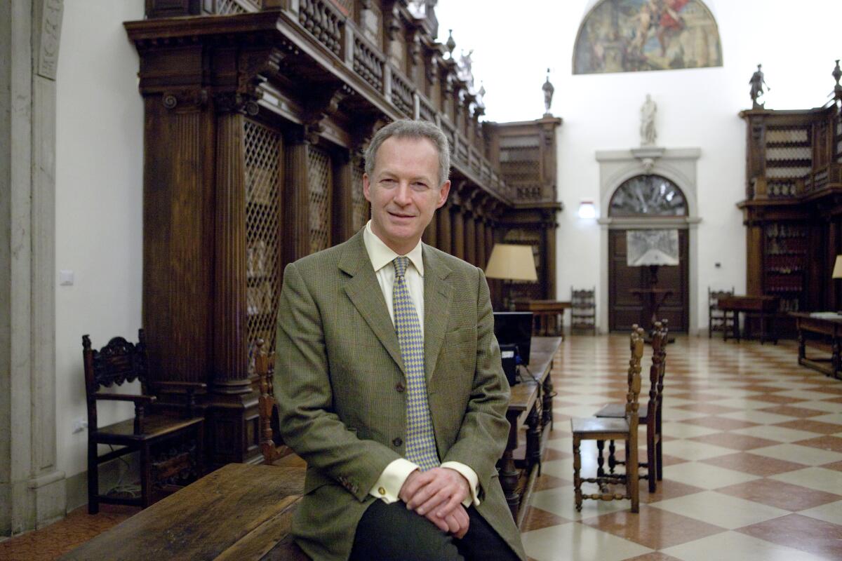 James Daunt sitting on a long wooden table in a historic building.