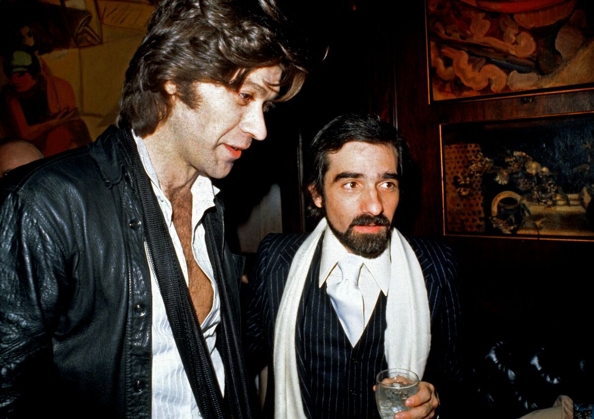 A man with a guitar and a filmmaker with a drink wearing a scarf pose for the camera