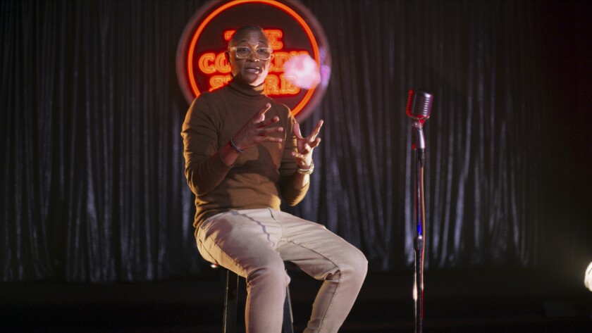 A man sits on a stool in front of a neon sign reading "The Comedy Store"