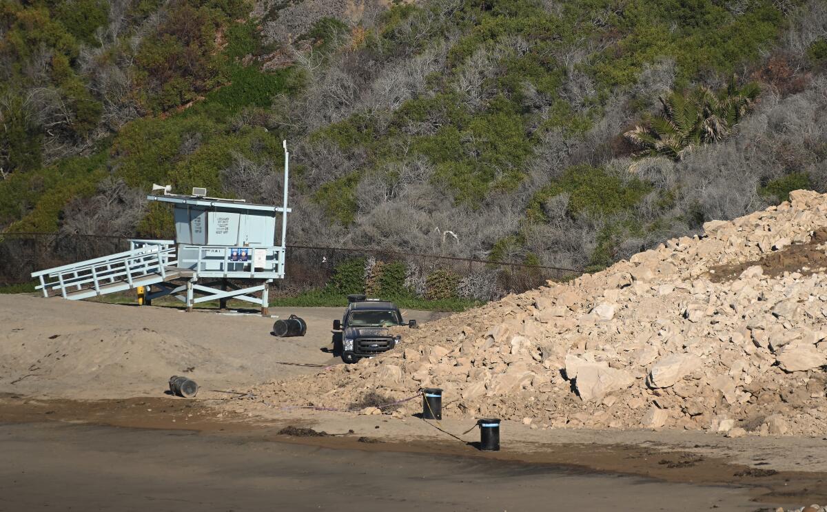 Rocky debris partially covers a pickup truck on a beach near a lifeguard tower