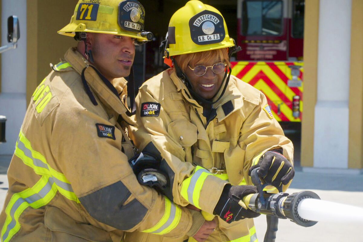A man and a woman in firefighter coats hold a fire hose that's spewing water.