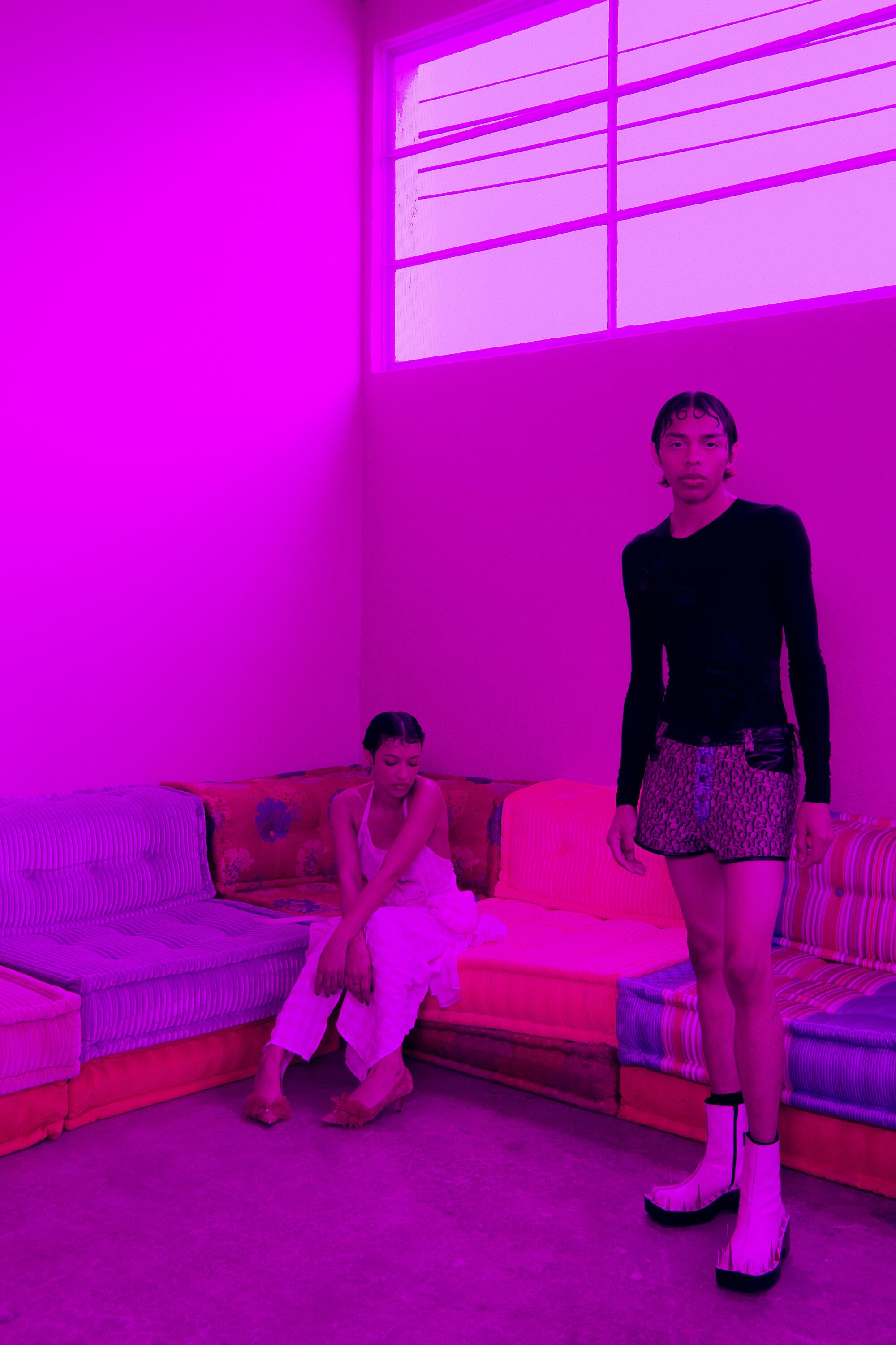 Two models bathed in a bright pink light, a colorful couch in the background.