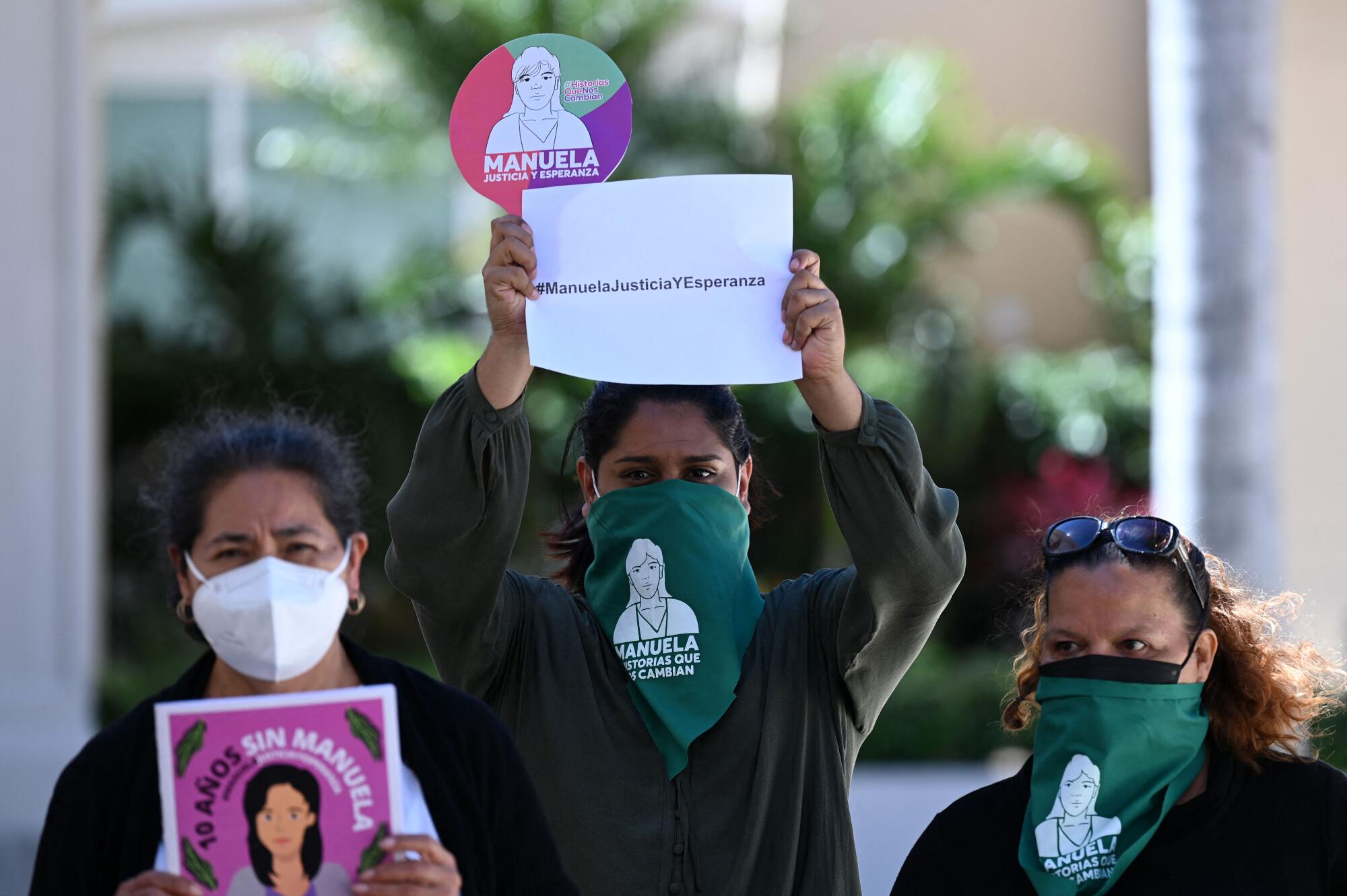 Women wearing green face coverings and a mask hold signs 