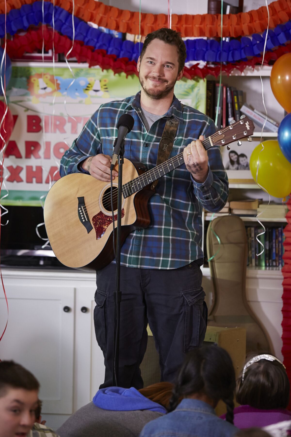 Chris Pratt holds a guitar behind a microphone in front of a birthday banner, with children seated on the ground.