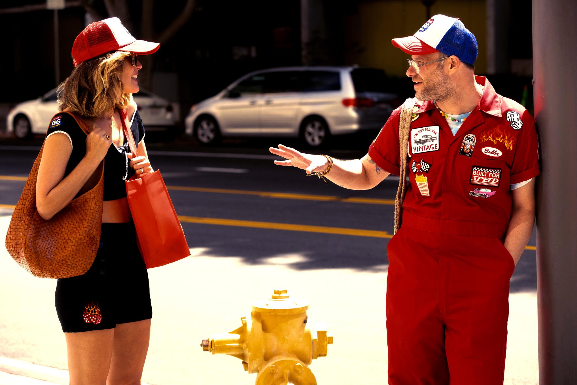 a man in a red jumpsuit, right, talks to a woman in shorts and a trucker hat carrying two bags 