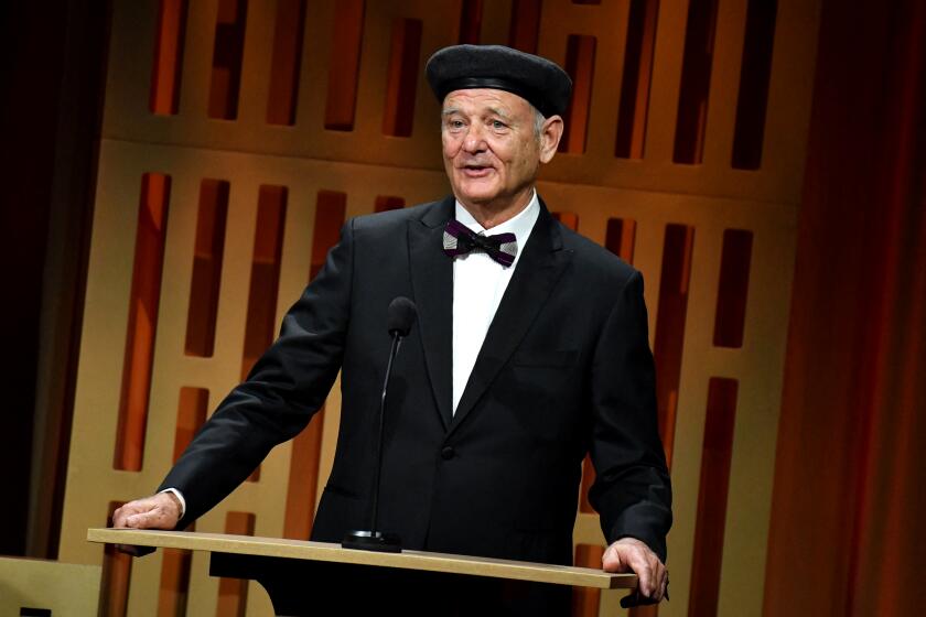 A man in a black hat and tuxedo speaking into a microphone on a stage